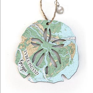 May include: A wooden sand dollar ornament with a nautical map of Tybee Island, Georgia. The ornament is painted in shades of blue and green and has a white pearl hanging from the top.