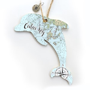 May include: Dolphin-shaped ornament featuring a nautical chart design with the text "Cedar Key." The ornament is light blue with green and white details, a compass rose tail, and a small compass charm. It hangs from a natural twine string.