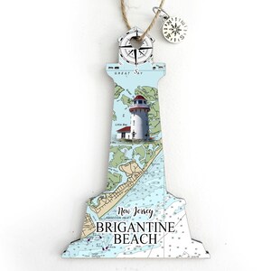 May include: A wooden lighthouse ornament with a nautical chart design featuring the text "New Jersey, Absecon Inlet, Brigantine Beach". The lighthouse has a red roof and a white tower with a small red building at the base. The ornament has a compass on top and a small compass charm hanging from the top.