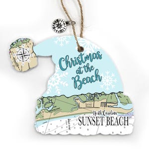 May include: A Santa hat-shaped ornament featuring a light blue top with white snowflakes and the text "Christmas at the Beach." The bottom section displays a nautical map of Sunset Beach, North Carolina, with a compass charm attached to the twine.