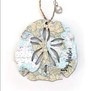 May include: A wooden sand dollar ornament with a nautical chart design and the text "Sag Harbor" printed on it. The ornament is white with blue and brown details and has a hole at the top for hanging.