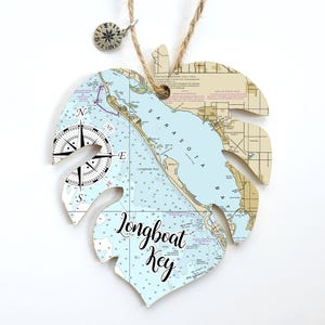 May include: A nautical map of Longboat Key, Florida, printed on a light blue and white monstera leaf-shaped ornament. The ornament has a compass rose and a twine hanger.