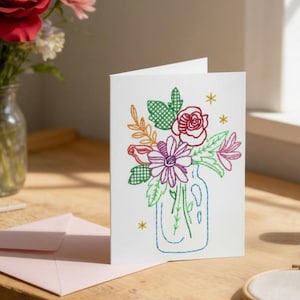 A handmade card featuring a colorful embroidered flower arrangement sits on a wooden table, accompanied by a vase of flowers, spools of thread, and a pair of scissors.