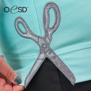 May include: A silver glitter scissors applique is displayed on a light turquoise fabric. The scissors are embroidered with a detailed design and are crossed. The OESD logo is in the upper left corner.