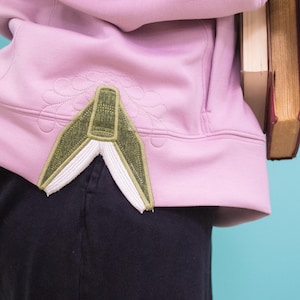 Puede incluir: Una sudadera con capucha rosa claro con un diseño bordado en verde y blanco. La sudadera se combina con pantalones azul oscuro. Dos libros se sostienen en la mano derecha.