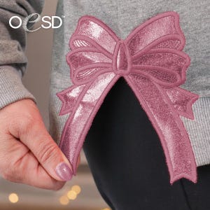 Peut inclure: Un sweat-shirt gris chiné orné d'une grande application de nœud brodé rose mauve métallique sur l'ourlet latéral. Le nœud présente des coutures détaillées et une texture scintillante. Le texte OESD est visible dans le coin supérieur gauche.