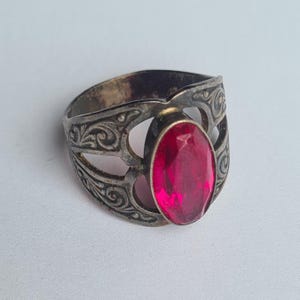 May include: A silver-toned metal ring featuring an oval-shaped, faceted red gemstone set in the center. The band displays an intricate, raised scrollwork pattern across its surface, with open cutouts surrounding the central stone setting.