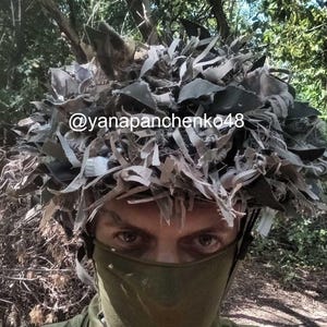 Peut inclure: Un couvre-casque de camouflage composé de bandes de tissu dans des tons vert olive, marron et gris, conçu pour la dissimulation. Le texte @yanapanchenko48 est imprimé sur le devant du couvre-casque, porté sur un masque facial vert olive uni.