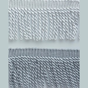 May include: Close-up of two decorative fringe trims. The top trim is white, and the bottom trim is a light grey. Both trims feature a twisted rope design with a woven header. Suitable for adding a decorative touch to clothing, home decor, or craft projects.