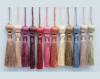 May include: A collection of decorative tassels in various colors, including brown, pink, red, and blue. Each tassel features a round, fabric-covered top and a long, fringed bottom. The tassels are suspended from thin cords, creating a visually appealing display.