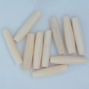 May include: A collection of eleven cream-colored, cylindrical wooden beads. Each bead has a rounded, tapered shape, with a smooth, unadorned surface. The beads are scattered on a light background.