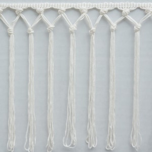 May include: Close-up of a white macrame fringe trim. The trim features a knotted top section with vertical strands of fringe hanging below. The fringe is made of a soft, white material, and the knots are evenly spaced.