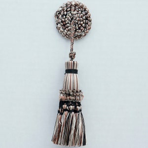 Peut inclure: Un pompon décoratif avec un motif rayé noir, blanc et marron. Le pompon a un dessus rond et un bas en frange. Le pompon est fait de tissu tissé.