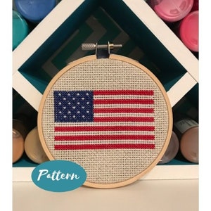 May include: A close-up of a finished cross-stitch embroidery project of the American flag in a wooden embroidery hoop. The flag is stitched in red, white, and blue thread on a beige fabric.