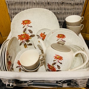 May include: A wicker picnic basket filled with a vintage floral tea set. The set includes a teapot, cups, saucers, and plates with a red floral pattern.