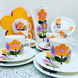 Op de afbeelding: Een vintage bloemen theeservies met een theepot, kopjes en schoteltjes. De set heeft een kleurrijk bloemenpatroon met roze, paarse en gele bloemen op een witte achtergrond.