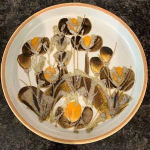 Può includere: Un piatto in ceramica bianca con un design floreale marrone e arancione. Il piatto ha un bordo marrone e un centro bianco con un motivo floreale in tonalità marroni e arancioni.