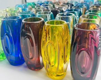May include: A collection of cylindrical glass vases in various vibrant colors including blue, yellow, purple, and green. Each vase features a smooth, polished surface with a distinct oval-shaped indentation on the side, creating a reflective effect.