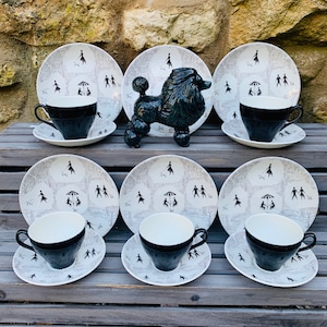 Puede incluir: Un juego de té vintage de mediados de siglo con seis platos, seis platillos y seis tazas de té negras. Los platos y platillos tienen un patrón gris y blanco con figuras. Una figura de caniche negro está en el centro.