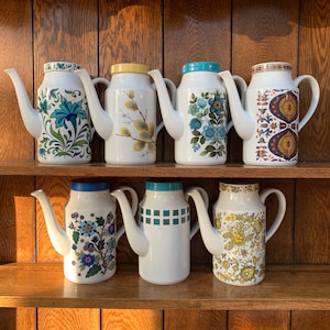 Può includere: Un set di sette caffettiere in ceramica vintage con motivi floreali e geometrici. Le caraffe sono bianche con accenti blu, verdi, gialli e marroni. Sono disposte su uno scaffale in legno.