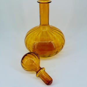 May include: Amber-colored glass decanter with a round body and a long neck. The decanter has a matching stopper. The glass has a ribbed design around the body. The decanter and stopper are on a white background.