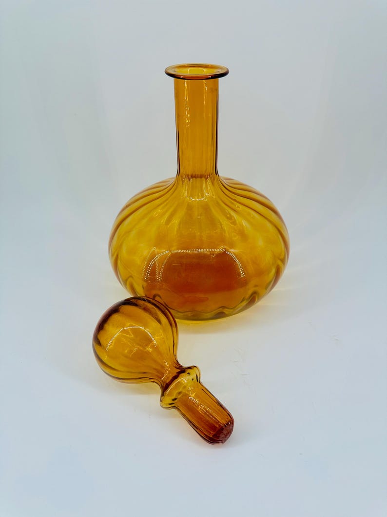 May include: Amber-colored glass decanter with a round body and a long neck. The decanter has a matching stopper. The glass has a ribbed design around the body. The decanter and stopper are on a white background.