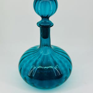 May include: A teal-colored glass decanter with a round body and a spherical stopper. The decanter has a ribbed design, and the stopper is also ribbed. The glass is transparent, allowing light to pass through. The decanter is likely used for storing and serving beverages.
