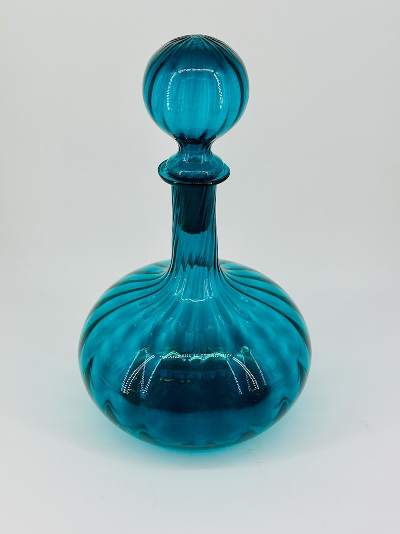 May include: A teal-colored glass decanter with a round body and a spherical stopper. The decanter has a ribbed design, and the stopper is also ribbed. The glass is transparent, allowing light to pass through. The decanter is likely used for storing and serving beverages.