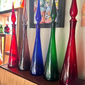 Op de afbeelding: Vijf hoge, slanke glazen karaffen met puntige stoppen, opgesteld in een rij. De kleuren omvatten helderrood, dieppaars, levendig blauw, smaragdgroen en een tweede helderrood, allemaal met gladde, conische lichamen en lange halzen.