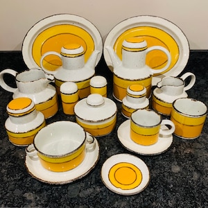 May include: A collection of vintage ceramic tableware with a white, yellow, and brown color scheme. The set includes teapots, serving platters, cups, saucers, sugar bowls, and creamers. The pieces have a speckled texture and a retro design.
