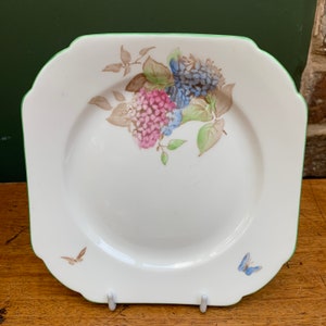 Op de afbeelding: Een wit vierkant bord met een groene rand heeft een bloemenontwerp met roze en blauwe bloemen met groene bladeren. Twee vlinders zijn op het bord geschilderd, een bruin en een blauw.