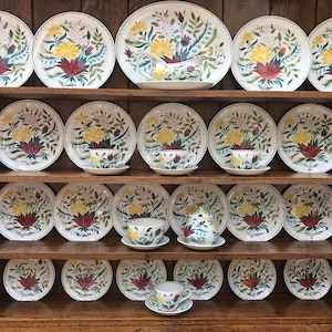 Puede incluir: Un juego de platos, cuencos y platillos de porcelana vintage con estampado floral. Los platos tienen un fondo blanco con un diseño floral colorido con flores rojas, amarillas y azules. El juego está dispuesto en un estante de madera.