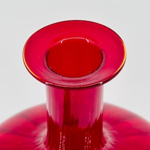 May include: A close-up of a vibrant red glass vase. The vase has a wide, circular opening with a thin, gold-colored rim. The body of the vase is a deep red, with a smooth, glossy finish. The background is plain white.