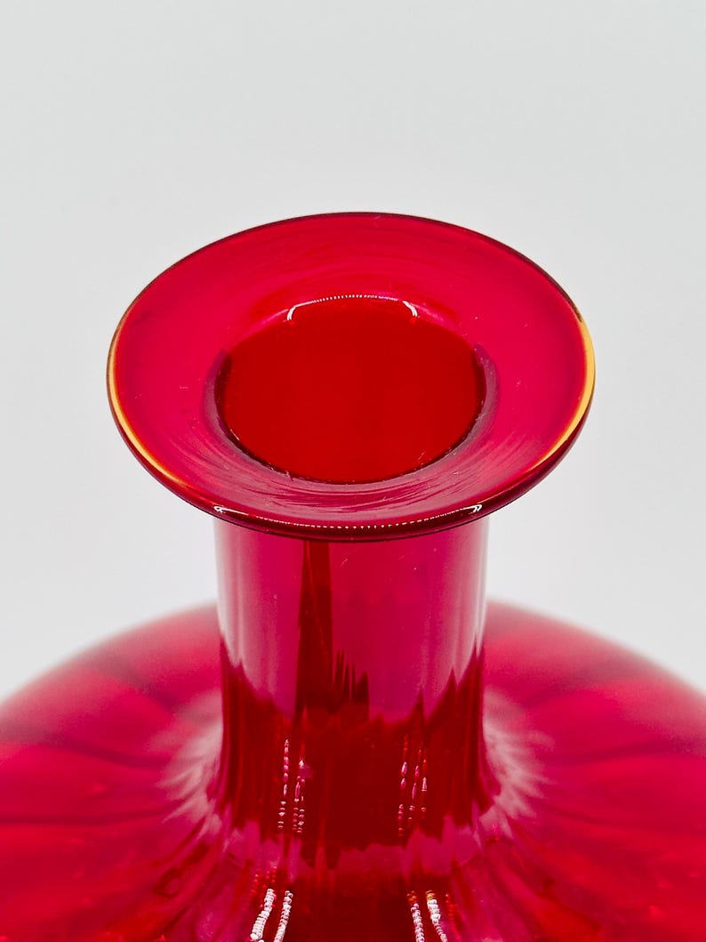 May include: A close-up of a vibrant red glass vase. The vase has a wide, circular opening with a thin, gold-colored rim. The body of the vase is a deep red, with a smooth, glossy finish. The background is plain white.