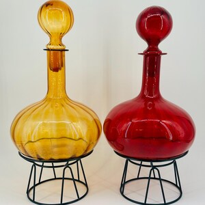 May include: Two decorative glass decanters, one amber and one red, each with a round body and a spherical stopper. They sit on black metal stands, showcasing a vintage aesthetic. The amber decanter has a ribbed design.