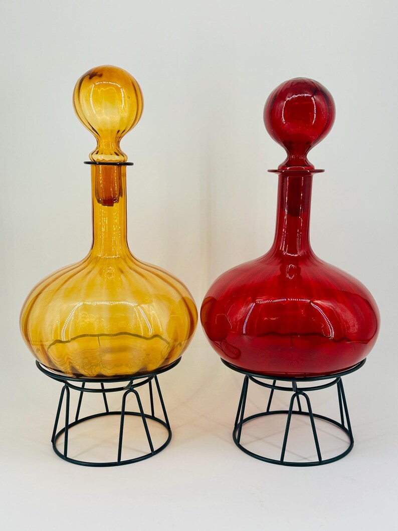 May include: Two decorative glass decanters, one amber and one red, each with a round body and a spherical stopper. They sit on black metal stands, showcasing a vintage aesthetic. The amber decanter has a ribbed design.