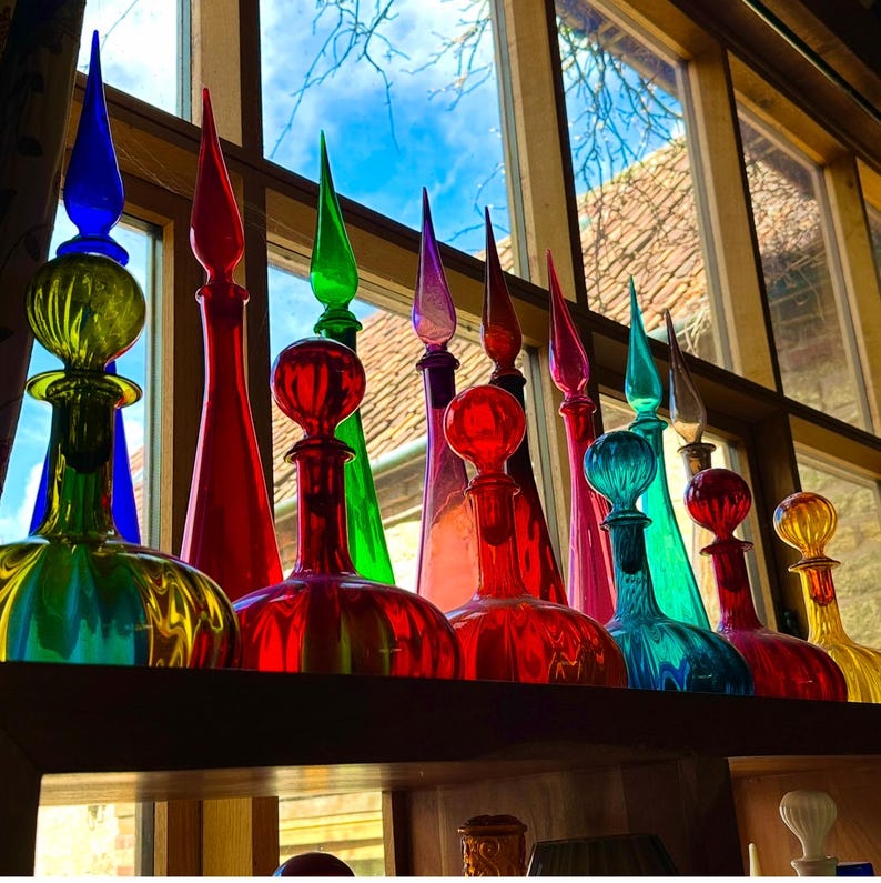 May include: A collection of colorful glass bottles with long, pointed stoppers. The bottles are in various colors, including red, green, blue, and yellow. They are arranged on a shelf in front of a window, with a view of the sky and a building.