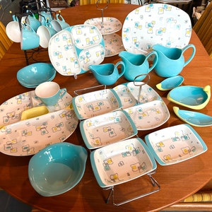 May include: A selection of vintage dishware featuring a cream base with a geometric pattern in yellow, blue, and grey. The set includes plates, bowls, cups, and serving dishes. Some pieces are a solid turquoise colour, creating a retro aesthetic.