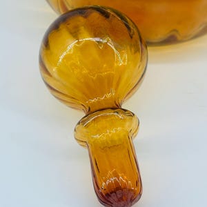 May include: Amber glass bottle stopper with a round, ribbed top and a flared base. The stopper is a warm amber color with darker brown accents. The background shows a portion of a matching amber glass bottle.