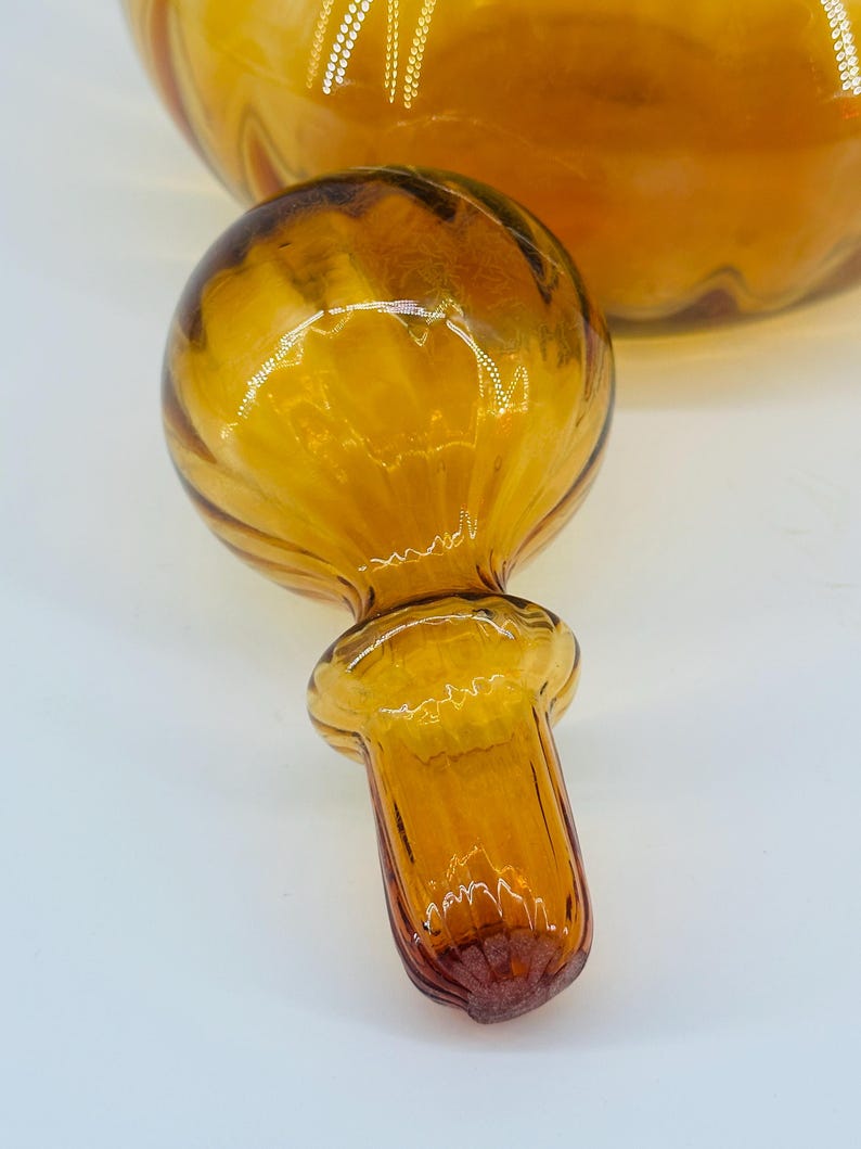 May include: Amber glass bottle stopper with a round, ribbed top and a flared base. The stopper is a warm amber color with darker brown accents. The background shows a portion of a matching amber glass bottle.