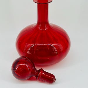 May include: A vibrant red glass decanter with a round body and a long neck. The matching stopper is lying next to the decanter. The glass has a glossy finish, reflecting light. The decanter is a decorative item.