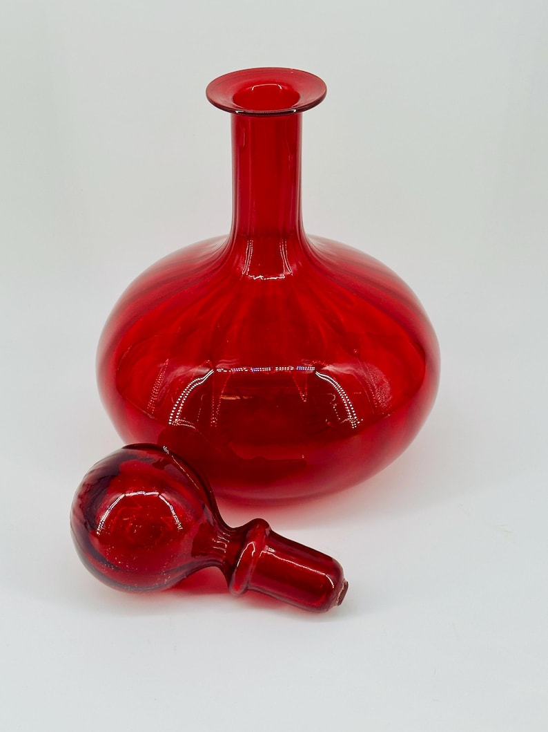 May include: A vibrant red glass decanter with a round body and a long neck. The matching stopper is lying next to the decanter. The glass has a glossy finish, reflecting light. The decanter is a decorative item.