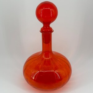 May include: A vibrant red glass decanter with a round, ribbed body and a long neck. The decanter is topped with a spherical stopper. The design is simple and elegant, suitable for serving wine or other beverages.