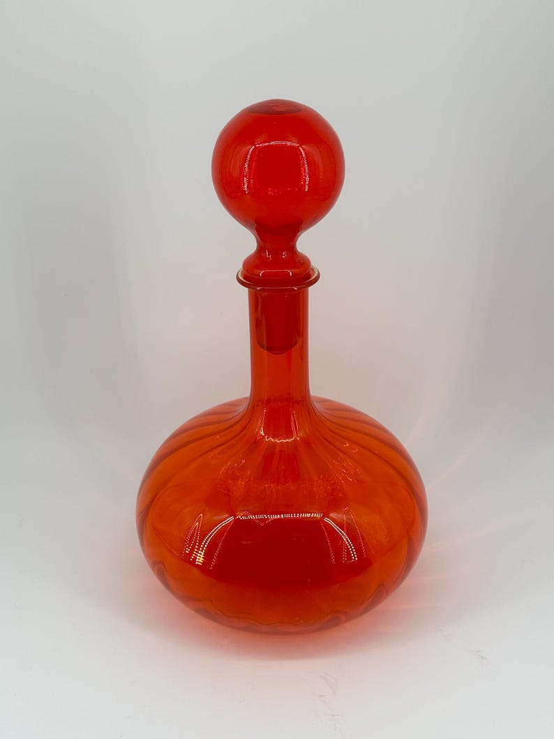 May include: A vibrant red glass decanter with a round, ribbed body and a long neck. The decanter is topped with a spherical stopper. The design is simple and elegant, suitable for serving wine or other beverages.