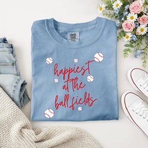 May include: A light blue t-shirt with the words "happiest at the ball fields" in red script, along with baseball graphics. The shirt is surrounded by a pair of white sneakers, blue jeans, and flowers.