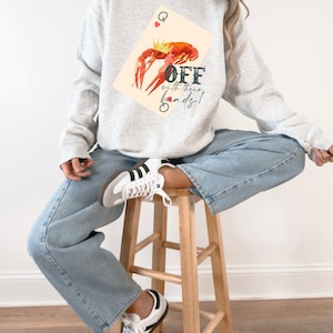 May include: Pale grey sweatshirt with a playing card design featuring a red crawfish wearing a gold crown. The card reads "Queen" and the text "OFF with their heads!" is below the crawfish. The sweatshirt is worn with light blue jeans.