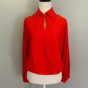 May include: A red long-sleeved blouse with a keyhole neckline and a button closure. The blouse has a collar and long sleeves with gathered cuffs.