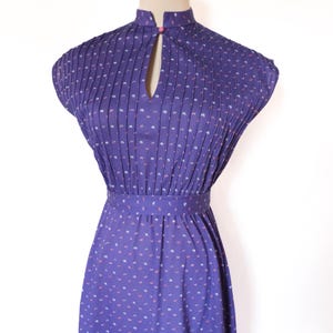 May include: A vintage purple dress with a high neckline and a keyhole detail. The dress features a pleated bodice and a fitted waist. It is adorned with a delicate pattern of small white and pink shapes. The dress is displayed on a mannequin.