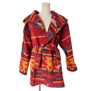 May include: A hooded robe with a geometric pattern in shades of red, yellow, blue, and white. The robe has a belt and is displayed on a mannequin. The robe's design features Aztec-inspired motifs.