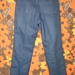 May include: Light blue denim jeans with a classic five-pocket design. The jeans have a straight leg cut and a Wrangler brand label on the back pocket. The jeans are displayed against a floral patterned background.